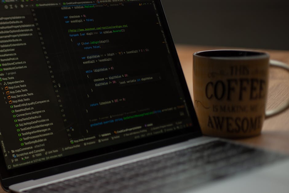 A close-up of a laptop displaying lines of green, blue, and white code on its screen, placed on a wooden surface. To the right of the laptop, there is a ceramic coffee mug with a humorous message that