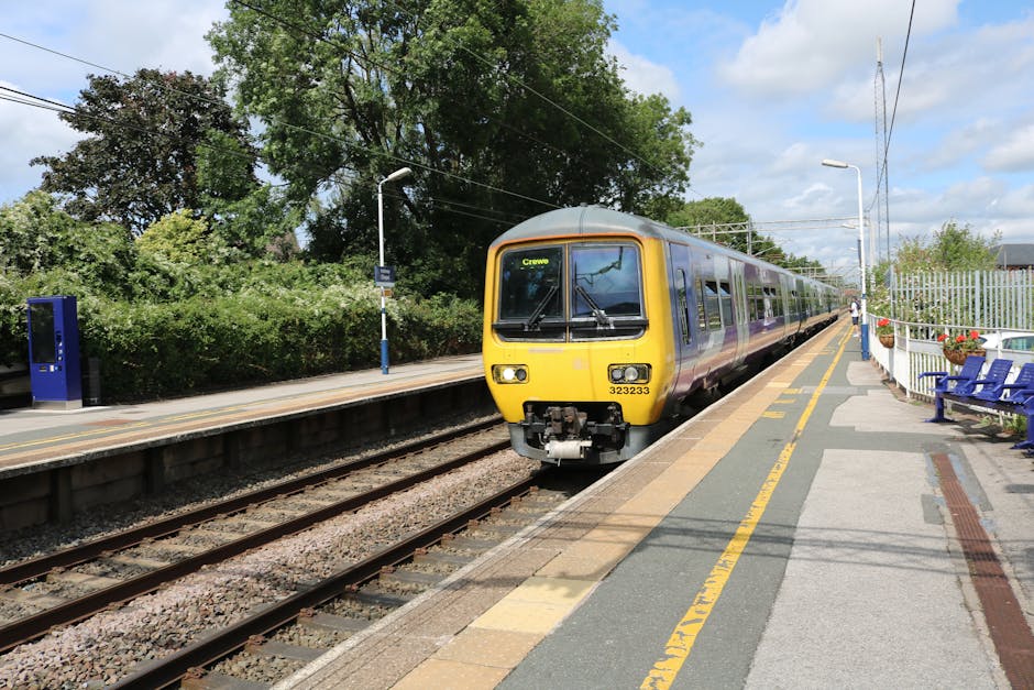 The image depicts a railway station platform with lush green trees and dense hedges in the background. A modern yellow and purple train with the destination display 'Crewe' is approaching or arriving 